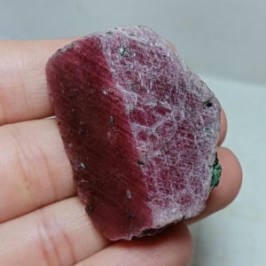 May include: A rough-cut ruby gemstone, held in a hand. The ruby displays a deep red hue with areas of lighter pink and white veining. Small black inclusions are visible throughout the stone, and a touch of green is seen on one edge.