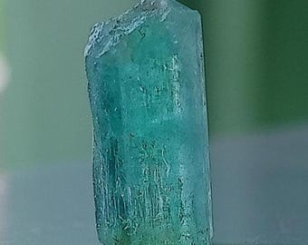May include: A raw, translucent crystal specimen with a vertical, elongated prismatic shape. The mineral displays a vibrant teal-green color with visible internal striations and a jagged, uneven top edge, showcasing a natural, unpolished texture.