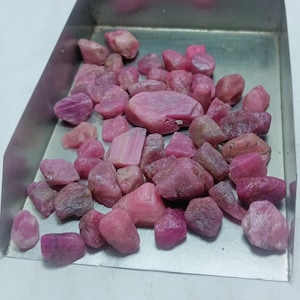 Puede incluir: Una colección de piedras de rubí rosa en bruto en una bandeja de metal. Las piedras son de varios tamaños y formas, algunas tienen un aspecto más pulido.
