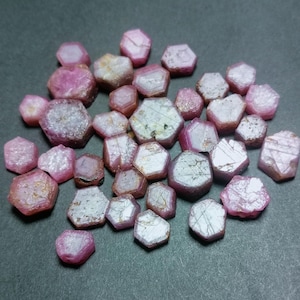 Op de afbeelding: Een verzameling roze robijnkristallen, elk met een zeshoekige vorm en een licht metalen glans. De kristallen zijn in een cluster op een donkergrijs oppervlak gerangschikt.