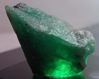 May include: A raw, uncut emerald gemstone. The gem is a deep, vibrant green color with a rough, textured surface. The emerald has a crystalline structure and a natural, organic shape, with a pointed tip.
