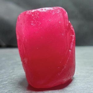 Puede incluir: Una pequeña piedra preciosa de color rosa vibrante y translúcido con una forma redondeada e irregular. La superficie de la piedra muestra una textura pulida y ligeramente desigual con sutiles facetas naturales y reflejos internos.