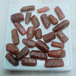 May include: A collection of rough, reddish-brown gemstones of varying sizes and shapes, scattered on a white tray. The stones have a natural, unpolished appearance, with visible textures and subtle color variations. The overall arrangement suggests a display of raw mineral specimens.