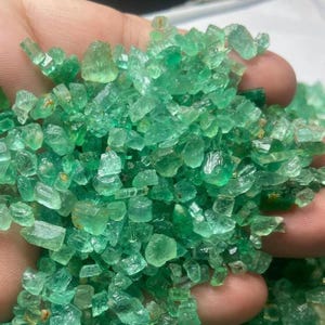 Può includere: Un pugno di cristalli di smeraldo grezzi. Le pietre preziose sono di un verde vibrante, con varie forme e dimensioni. I cristalli hanno una qualità traslucida, catturando la luce.