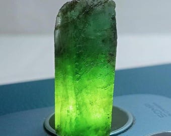 Puede incluir: Un cristal verde crudo y translúcido con una superficie rugosa y texturizada. El cristal está iluminado desde dentro, creando un brillo vibrante. La piedra preciosa tiene forma rectangular, mostrando formaciones minerales naturales.