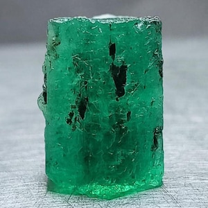 May include: A raw, hexagonal emerald crystal with a vibrant green color and a translucent, textured surface. The gemstone features natural inclusions and dark, irregular mineral deposits scattered throughout its vertical, prismatic structure.