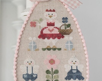 May include: A cross-stitch design of a chick in a red dress holding a basket of flowers, two chicks in overalls, and a potted flower. The design is stitched on a light brown fabric with a pink pom-pom trim.