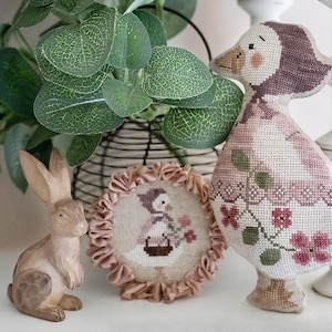 Può includere: Un'esposizione decorativa con un coniglietto in ceramica, un ricamo a punto croce incorniciato e un'oca di stoffa. L'oca e il ricamo presentano motivi floreali e geometrici in rosa e verde. Il coniglietto è marrone chiaro.