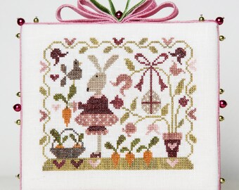 May include: A white cross-stitch embroidery with a pink border and gold beads. The design features a bunny holding a carrot in a garden setting with flowers, birds, and a basket of carrots.