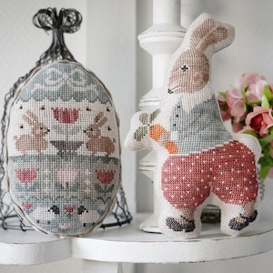 May include: A decorative Easter-themed display featuring two cross-stitched fabric figures. One is an egg-shaped ornament with bunnies and floral patterns. The other is a bunny holding a carrot, wearing a blue vest and red polka-dot pants.