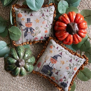 May include: Two embroidered pillow covers with a fall theme. The pillows are beige with brown trim and feature a pumpkin-faced character with a hat and a broom. The pillows are surrounded by green leaves and pumpkins.