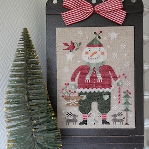 Peut inclure: Une broderie au point de croix encadrée représentant un bonhomme de neige en veste rouge, pantalon vert et chapeau vert. Le bonhomme de neige tient un panier de fleurs et une canne en bonbon. Un nœud à carreaux rouges et blancs orne le haut du cadre. Un petit sapin de Noël décoratif est à gauche.