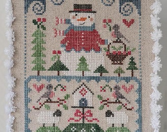 May include: A cross-stitch pattern depicting a snowman wearing a red hat and scarf, two birds carrying a basket, a birdhouse, and two sheep. The background is a blue sky with snowflakes.