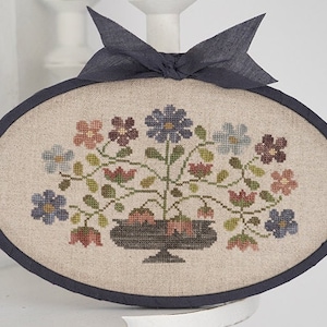 May include: A hand-stitched embroidery hoop with a floral design. The hoop is oval shaped and has a dark blue ribbon tied around it. The embroidery features a variety of flowers in shades of blue, pink, and purple, arranged in a vase.