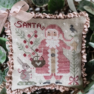 Peut inclure: Un coussin brodé représentant le Père Noël, un lapin et le mot "SANTA". Le Père Noël porte un costume rouge et tient un panier de baies. Le coussin a un bord à volants et un ruban rose.