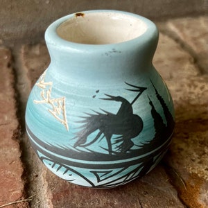 May include: A small, light blue ceramic vase with a black and white design of a bird with a spear. The vase has a rounded bottom and a wide opening.