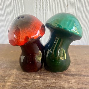 May include: A pair of ceramic mushroom-shaped salt and pepper shakers. One shaker is red and brown, the other is green. The shakers have a glossy finish and are designed to resemble mushrooms. The salt shaker has three holes on top.