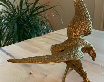 May include: A gold-colored brass eagle sculpture with its wings spread. The eagle is perched on a small branch and has a detailed, textured surface.