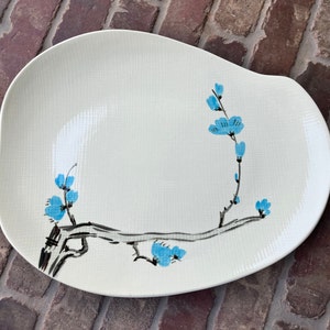 May include: A white ceramic serving platter with a blue and black floral design. The design features a stylized branch with blue flowers and black leaves.