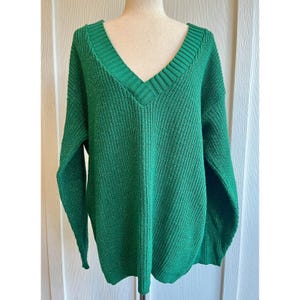 May include: A green, oversized V-neck sweater. The sweater has a ribbed neckline and cuffs, and a loose, comfortable fit. The fabric appears to be a chunky knit, perfect for cooler weather. The sweater is displayed on a mannequin.