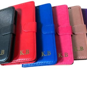 May include: A collection of colorful phone cases in black, red, blue, pink, tan, and purple. Each case is a folio style with a magnetic closure and features the gold initials "K.B."