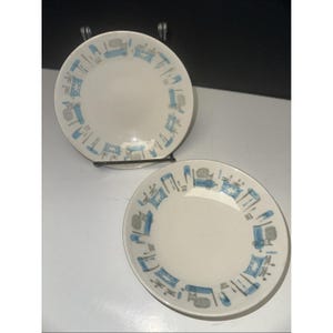 May include: Two white ceramic plates with a blue and gray furniture pattern around the rim. The plates are displayed on a metal stand against a dark background. The plates are likely for serving food.