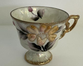 May include: A white teacup with a gold handle and base, adorned with a floral design in gold, pink, and black. The cup has a pedestal base and a gold rim, creating an elegant aesthetic.