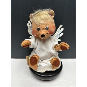 May include: A plush angel bear with blonde hair, a white robe, and wings. The bear is sitting on a black base.