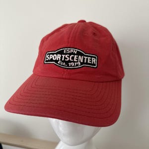 May include: Red baseball cap with a curved brim. The cap features the ESPN SportsCenter logo embroidered in white, black, and red. The text "ESPN SPORTSCENTER Est. 1979" is displayed on the front of the cap.