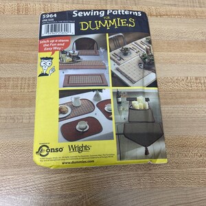 May include: Sewing pattern for table runners and placemats from the book "Sewing Patterns for Dummies". The pattern includes four different designs, all featuring a plaid pattern in red, white, and black. The pattern is printed on a yellow background with the title "Sewing Patterns for Dummies" in black text.