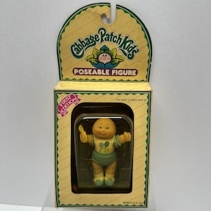 Peut inclure: Une figurine articulée Cabbage Patch Kids vintage dans son emballage d'origine. La figurine est un bébé avec un teint jaune, portant une couche et un bavoir verts. L'emballage est jaune avec les mots "Cabbage Patch Kids" et "Poseable Figure".