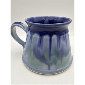 May include: A ceramic mug featuring a glossy glaze with a deep blue rim that transitions into a lighter periwinkle and muted green base. The mug has a wide, flared body and a curved handle on the left side, showcasing a smooth, drip-like finish.