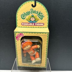 Puede incluir: Figura articulada Mimi Kara de Cabbage Patch Kids en su embalaje original. La muñeca tiene pelo naranja y viste un conjunto naranja con un estampado de rombos. La caja es amarilla y verde con las palabras "POSEABLE FIGURE" y "FIRST EDITION".