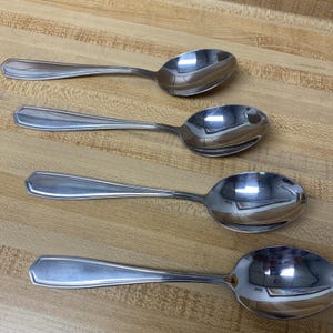 May include: Four shiny, silver spoons with a classic design are arranged on a light brown wooden surface. Each spoon has a rounded bowl and a handle with a subtle, geometric shape. The spoons reflect the light, highlighting their polished finish.