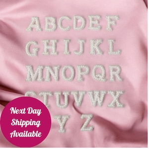 May include: Alphabet letters A through Z, each crafted with a pearl-like material, are displayed on a pink satin background. The letters are arranged in rows, showcasing the full alphabet. The image also includes a circular graphic with the text "Next Day Shipping Available."