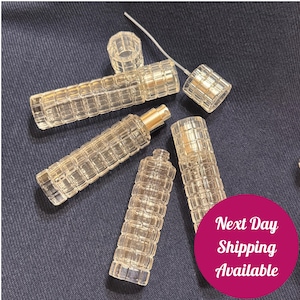May include: Several clear, textured glass perfume bottles and caps are arranged on a dark blue surface. The bottles have a rectangular grid pattern. One bottle has a gold-colored atomizer. The image includes a pink circle with the text "Next Day Shipping Available."