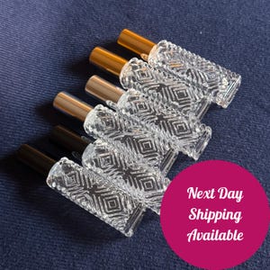 May include: Six clear glass perfume bottles with geometric diamond patterns. The bottles have gold, silver, and black colored caps. The bottles are arranged on a dark blue denim surface. The image also contains the text "Next Day Shipping Available".