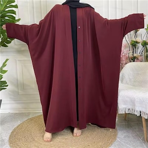May include: A burgundy abaya with a buttoned front and wide sleeves. It is worn over a black garment. The model is standing on a round woven mat. The abaya is a modest garment.