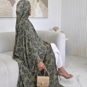 Puede incluir: Abaya verde oliva con estampado floral y velo a juego. El abaya se combina con pantalones blancos y tacones nude. Se sostiene en la mano un pequeño bolso beige con cierre dorado.