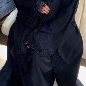 May include: A dark navy blue abaya featuring intricate floral lace trim along the front opening and sleeve cuffs. The garment is made of a smooth, solid fabric and includes delicate embroidery details integrated into the lace borders.
