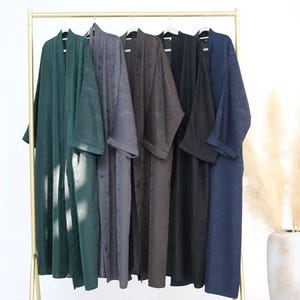 May include: A selection of long robes in shades of green, grey, and navy blue hang on a clothes rail. Each robe features a subtle floral design and wide sleeves. The robes are suspended from white hangers on a gold-coloured rail.