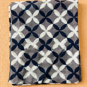 May include: Folded gray fabric with a repeating pattern of white and navy blue quatrefoils. The fabric has a textured appearance and is likely cotton or a similar material. The design is symmetrical and creates a visually appealing textile.