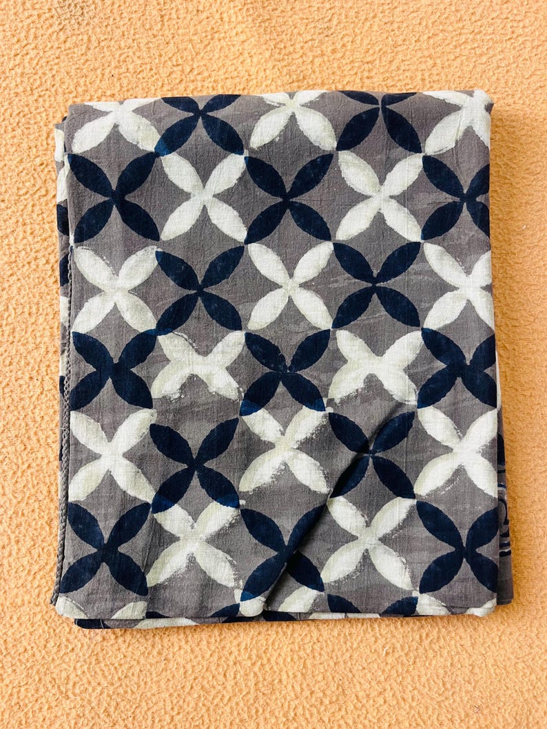 May include: Folded gray fabric with a repeating pattern of white and navy blue quatrefoils. The fabric has a textured appearance and is likely cotton or a similar material. The design is symmetrical and creates a visually appealing textile.