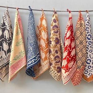 May include: A display of nine patterned cotton napkins hanging from a line. The napkins showcase diverse designs, including floral, animal, and geometric patterns in shades of red, blue, orange, and black. Each napkin is approximately 45 cm square.