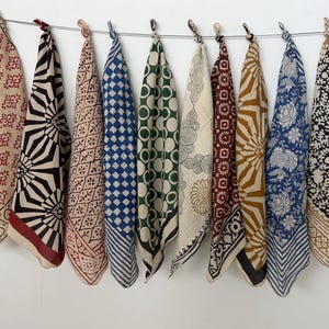May include: Ten square fabric cloths hanging from a wire, each featuring a unique block-printed pattern in colours including red, black, blue, green, and mustard yellow on cream-coloured backgrounds, with various geometric, floral, and circular motifs.