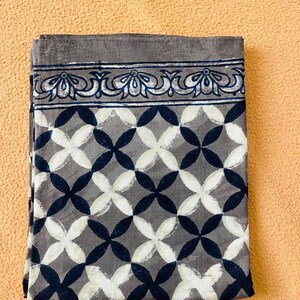 May include: Folded gray fabric with a geometric pattern of white and navy blue overlapping circles. The top border features a decorative floral design in white and navy blue. The fabric is displayed on a peach-colored surface.