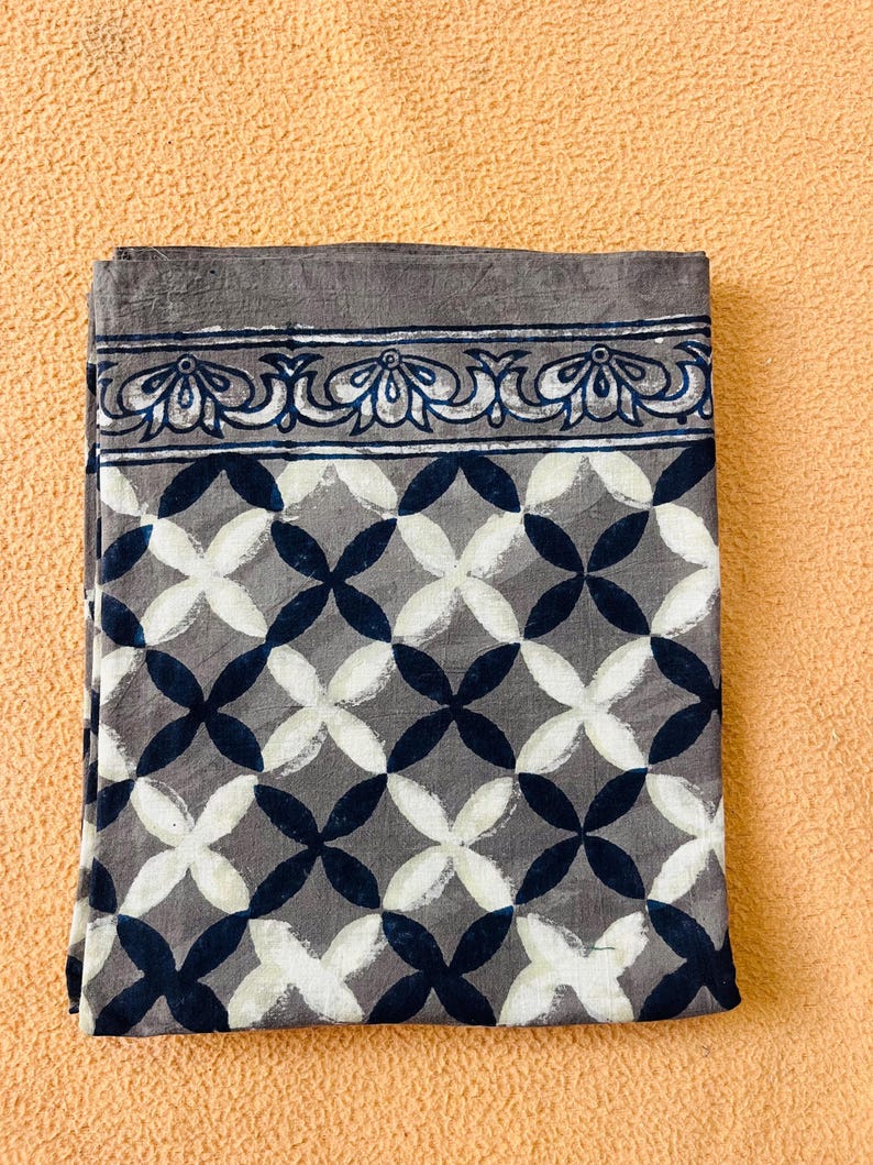 May include: Folded gray fabric with a geometric pattern of white and navy blue overlapping circles. The top border features a decorative floral design in white and navy blue. The fabric is displayed on a peach-colored surface.
