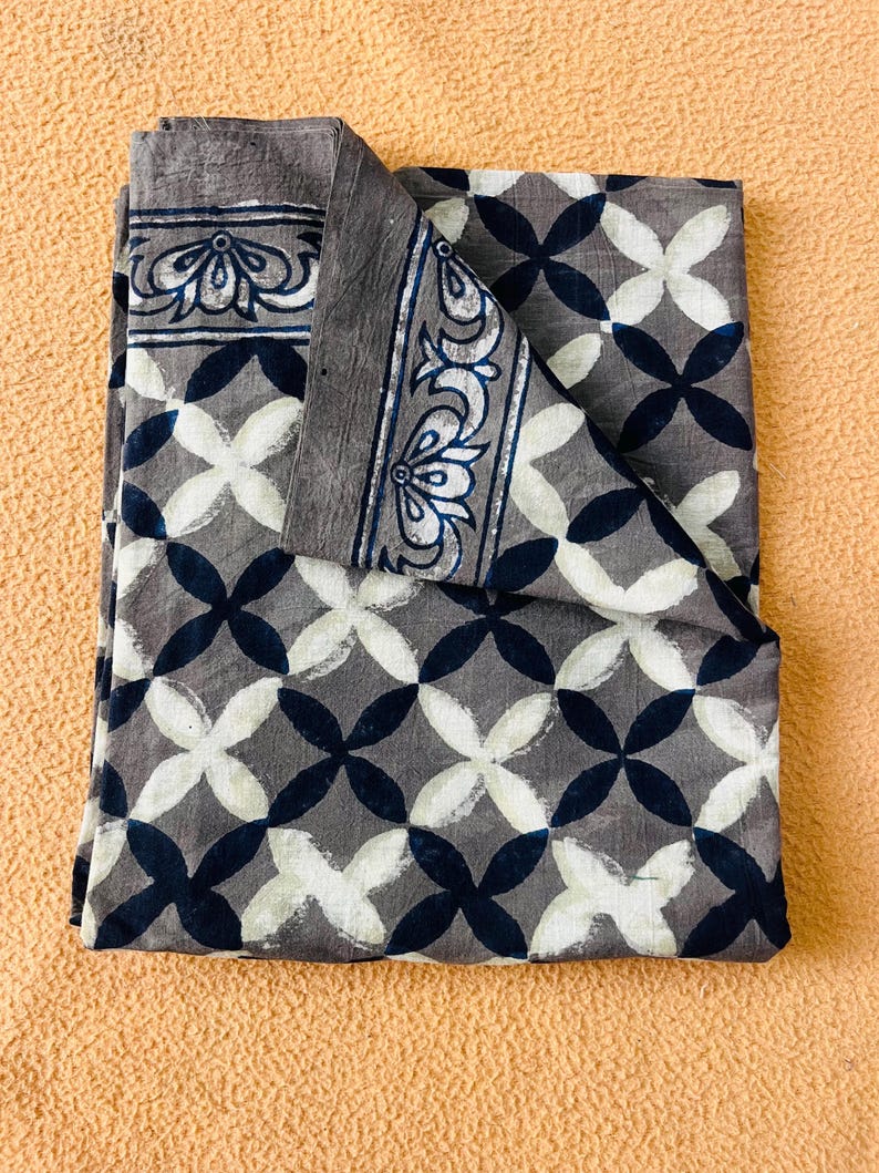 May include: Folded gray fabric with a repeating pattern of white and navy blue overlapping four-petal shapes. The border features a decorative floral design in white and navy blue.