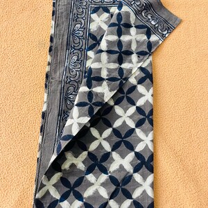 May include: Folded gray fabric with a geometric pattern of white and navy blue quatrefoils. The border features a decorative floral design in navy blue. The fabric is folded over, revealing the pattern and border details.