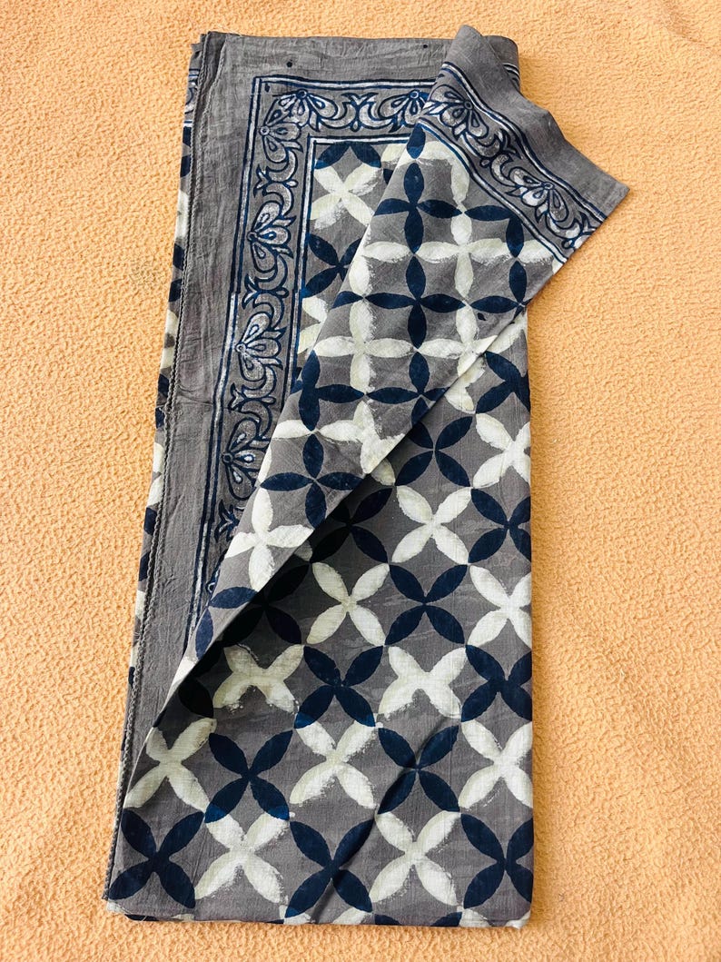 May include: Folded gray fabric with a geometric pattern of white and navy blue quatrefoils. The border features a decorative floral design in navy blue. The fabric is folded over, revealing the pattern and border details.